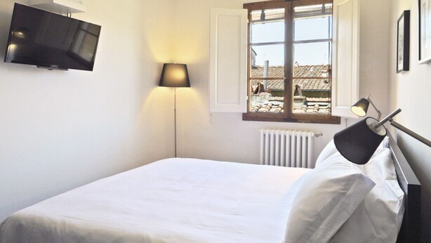 Un dormitorio con cama, televisor montado en la pared, ventana con vista a edificios y un radiador.