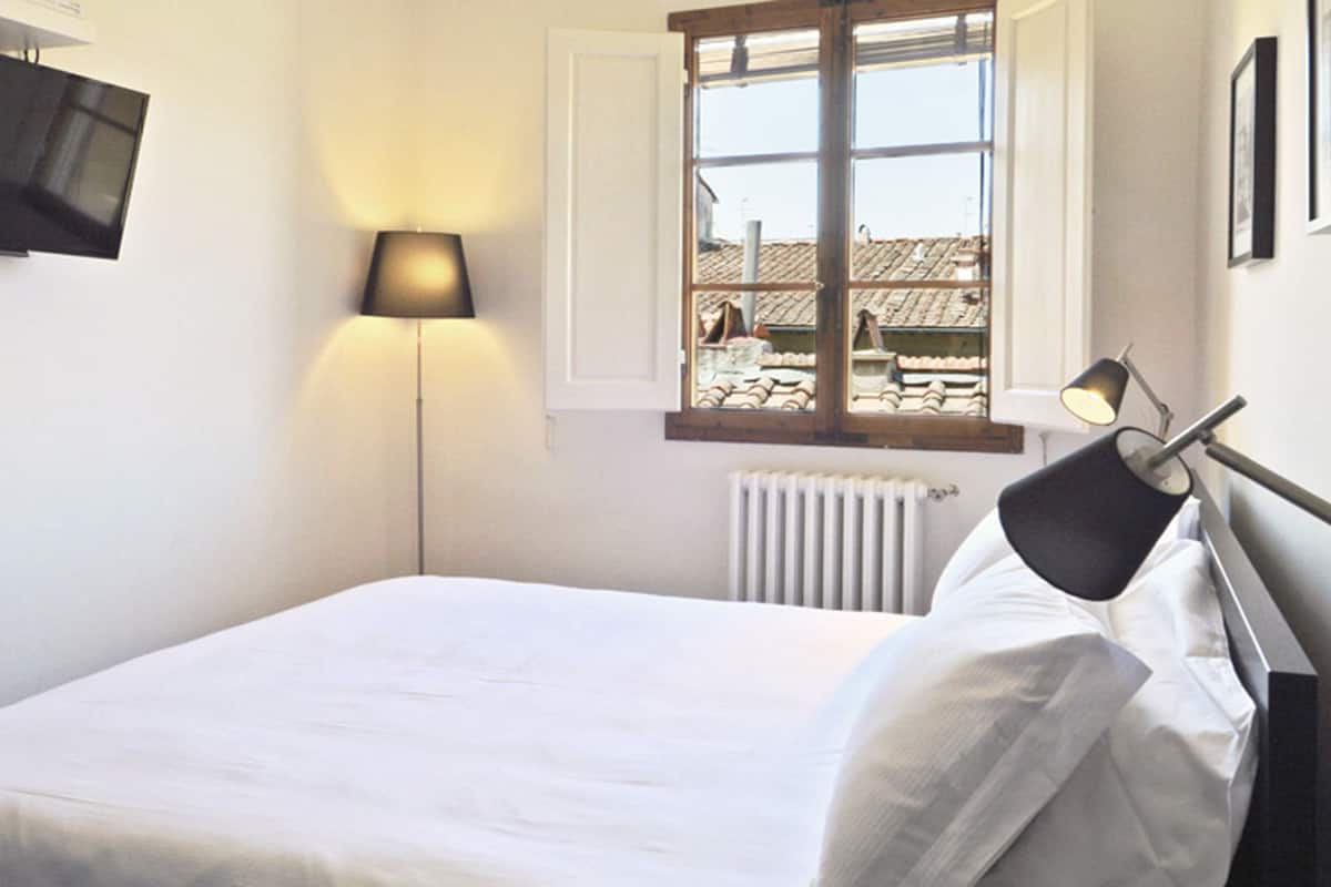 A bedroom with a bed, a TV mounted on the wall, a window with a view of buildings, and a radiator.