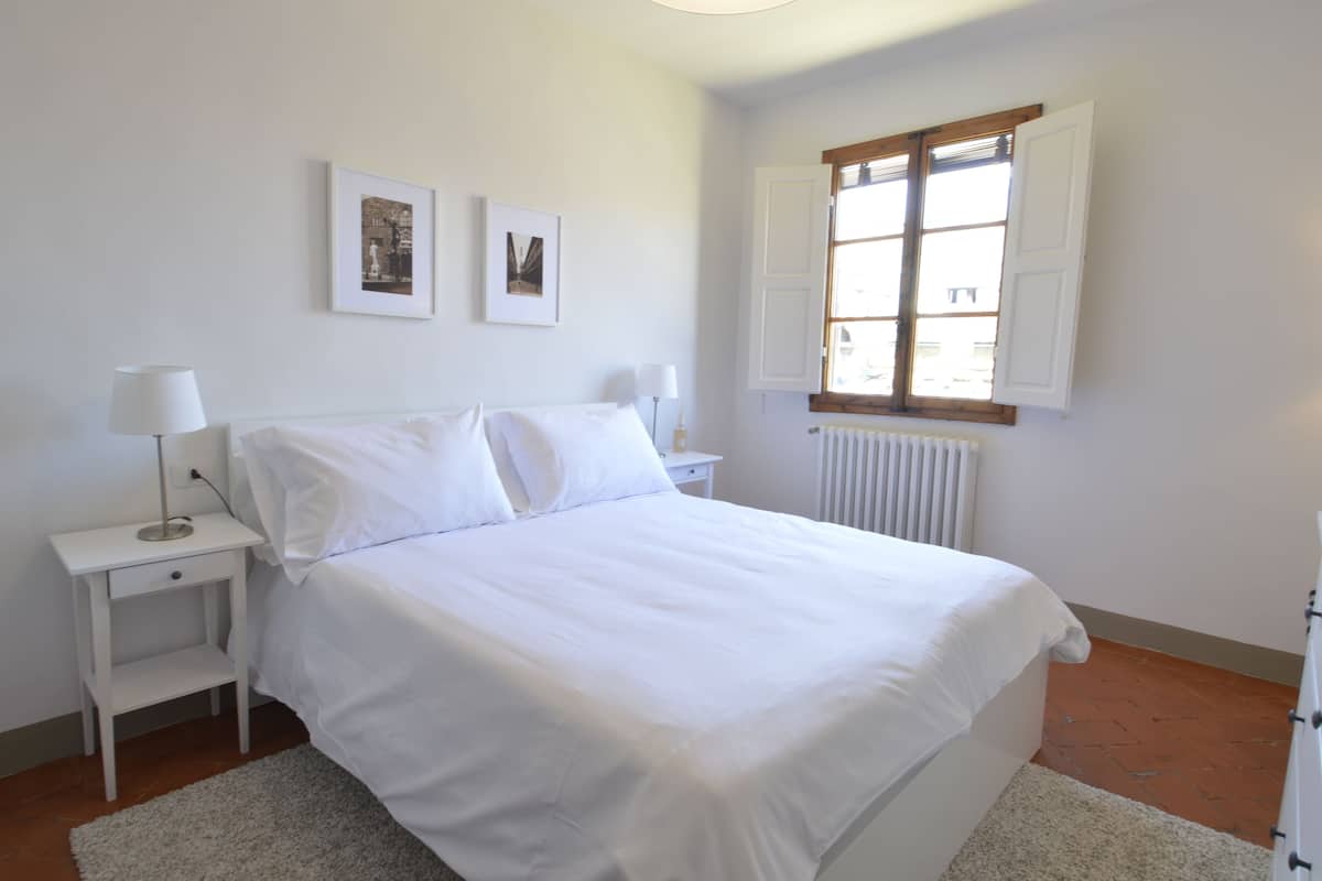 A white bedroom with a bed, two bedside tables, a window, and two framed pictures on the wall.