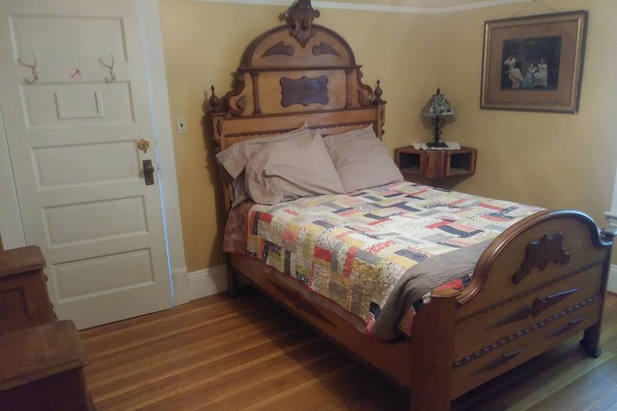 A wooden bed with a quilt, a nightstand with a lamp, and a framed picture on the wall.