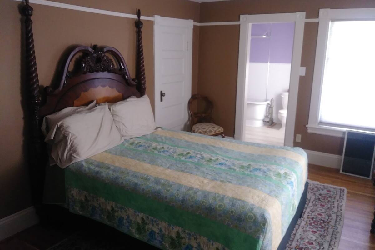 A bedroom with a four-poster bed, a wooden headboard, and a floral-patterned bedspread. There is a chair in the corner and a window with a view of the outside.