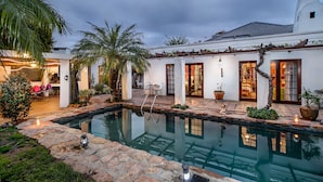 A pool area with a stone border, palm trees, and a building with white walls and wooden doors.