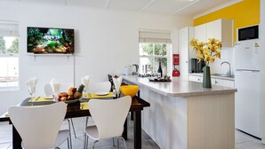 A modern kitchen with white cabinets, a yellow accent wall, a flat-screen TV mounted above the counter, and a dining area with white chairs and a fruit bowl.