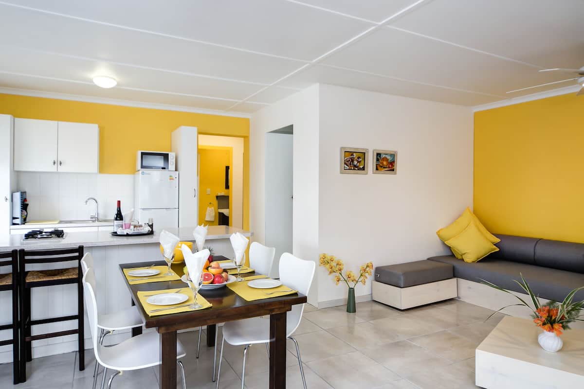 A modern kitchen with white cabinets, a dining area with a wooden table, and a living area with a sofa and yellow walls.