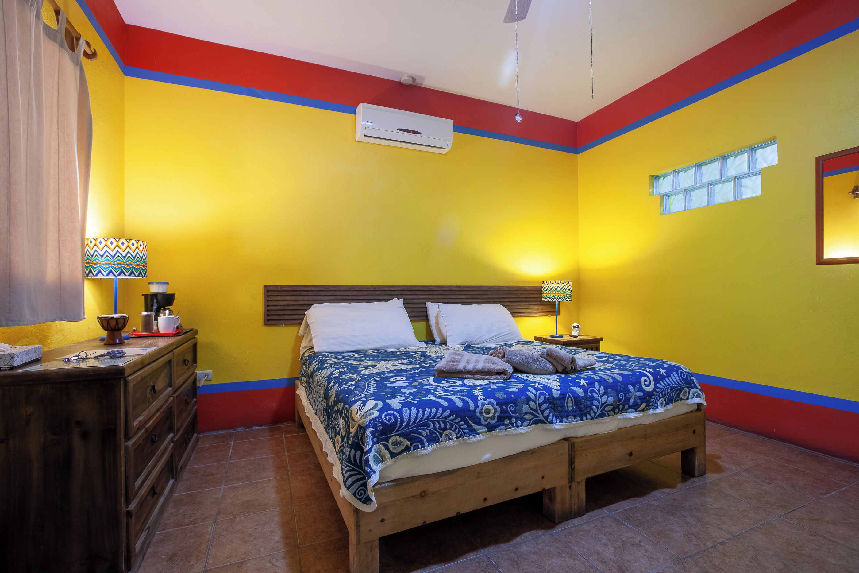 Un dormitorio con cama, mesitas de noche, cómoda, unidad de aire acondicionado, ventana y ventilador de techo.