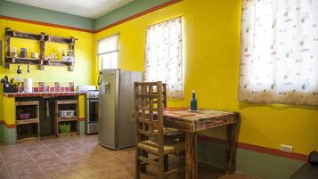 A kitchen with yellow walls, a wooden table, and a chair. There is a refrigerator, a sink, and a window with curtains.