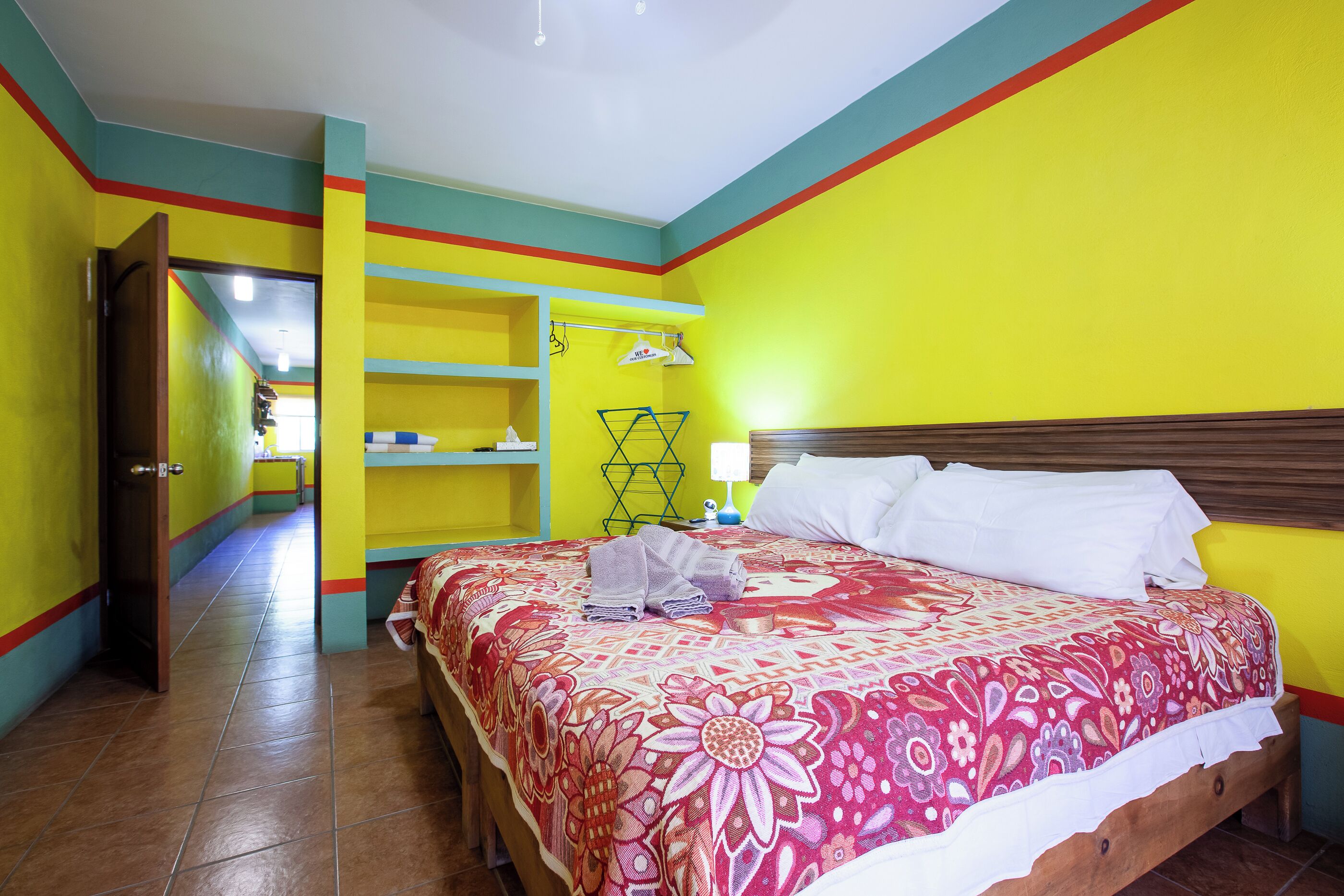 A brightly colored hotel room with a bed, a nightstand, a shelf with toiletries, and a door leading to another room.