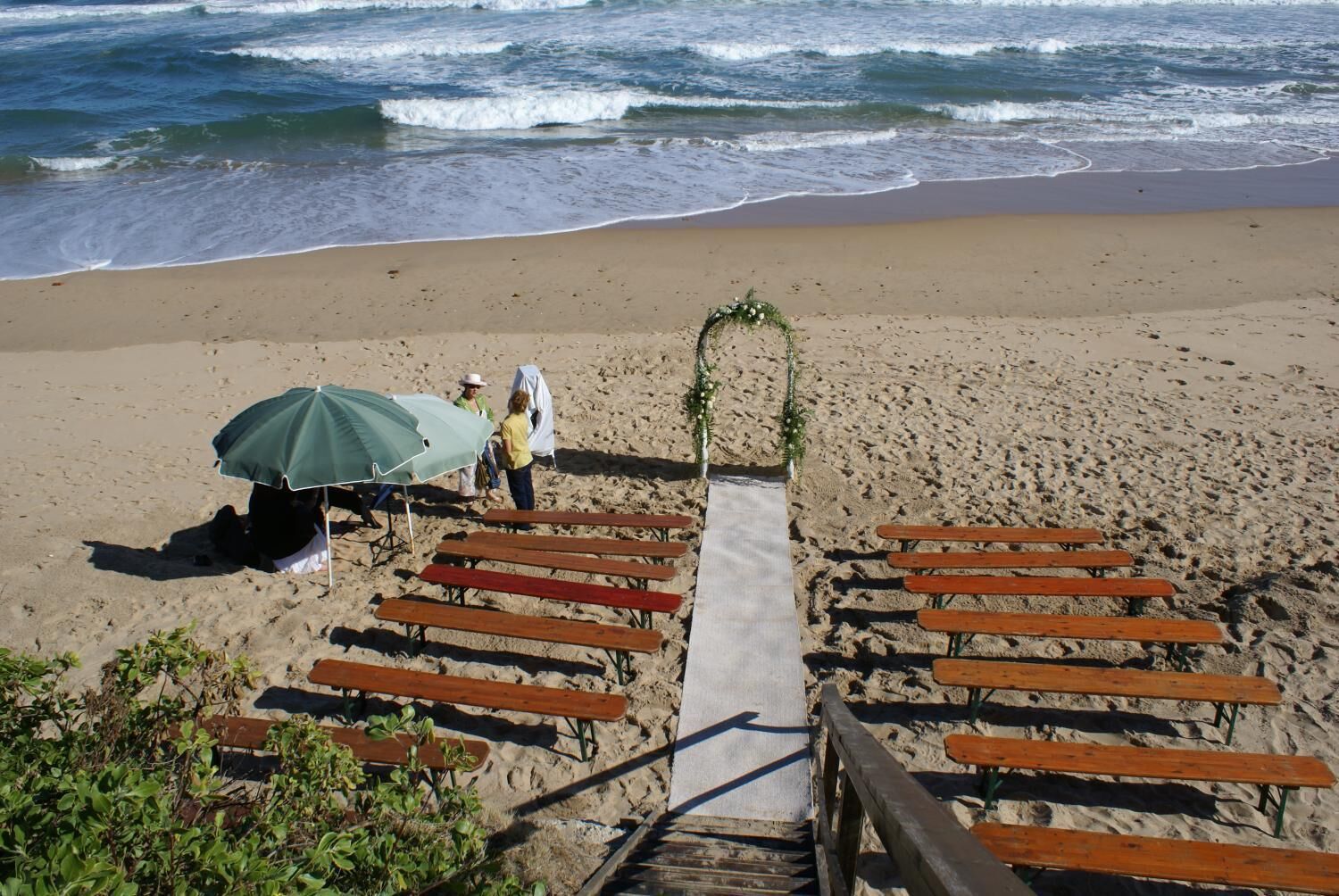 Decoración para una boda en la playa, con un sendero que lleva a un arco adornado, bancos de madera y un área techada con sombrilla.