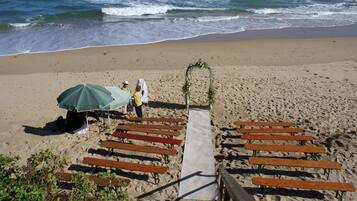 Decoración para una boda en la playa, con un sendero que lleva a un arco adornado, bancos de madera y un área techada con sombrilla.