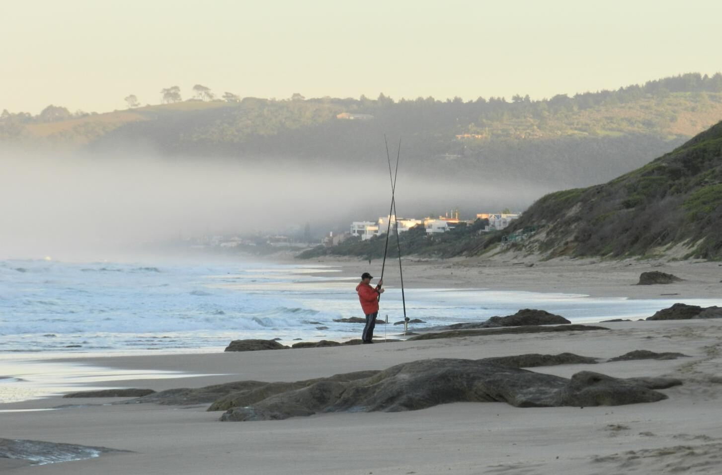 Une scène de plage avec une personne qui pêche, un rivage rocheux et des bâtiments en arrière-plan.