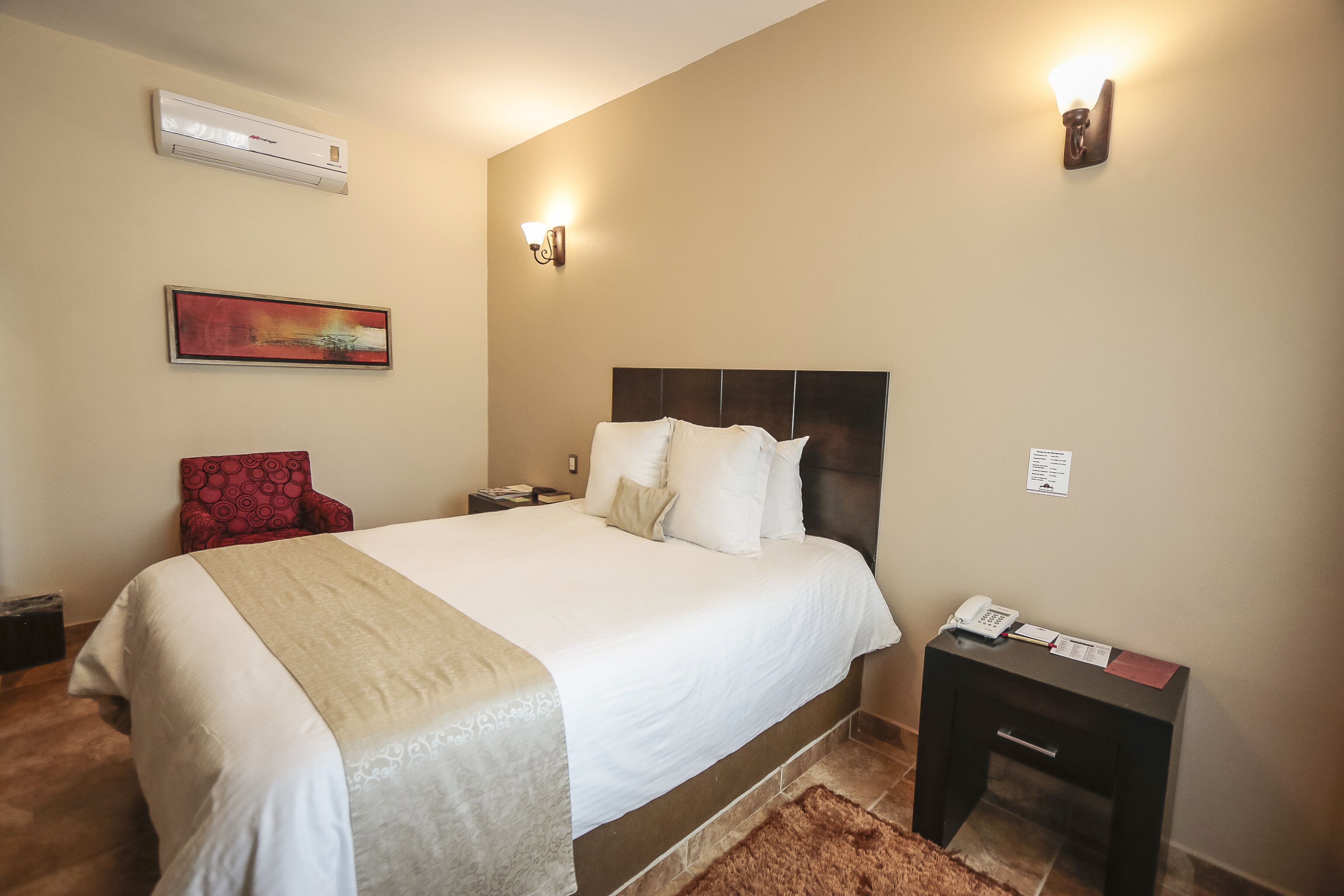 A hotel room with a large bed, a nightstand with a phone, a red chair, and a painting on the wall.