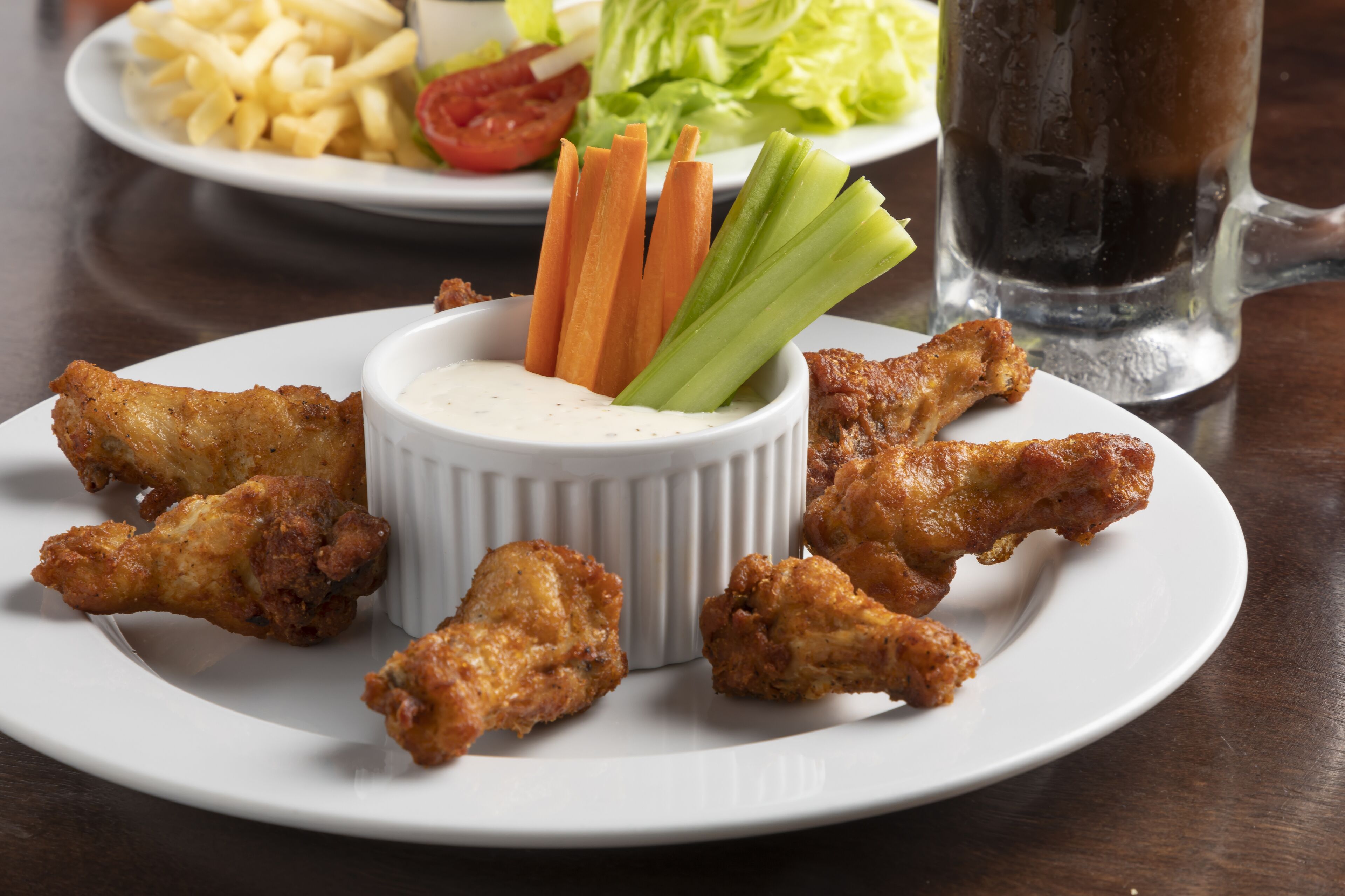 Un plato de alitas de pollo fritas con apio y zanahorias, un recipiente pequeño para salsa y un vaso de refresco sobre una mesa.