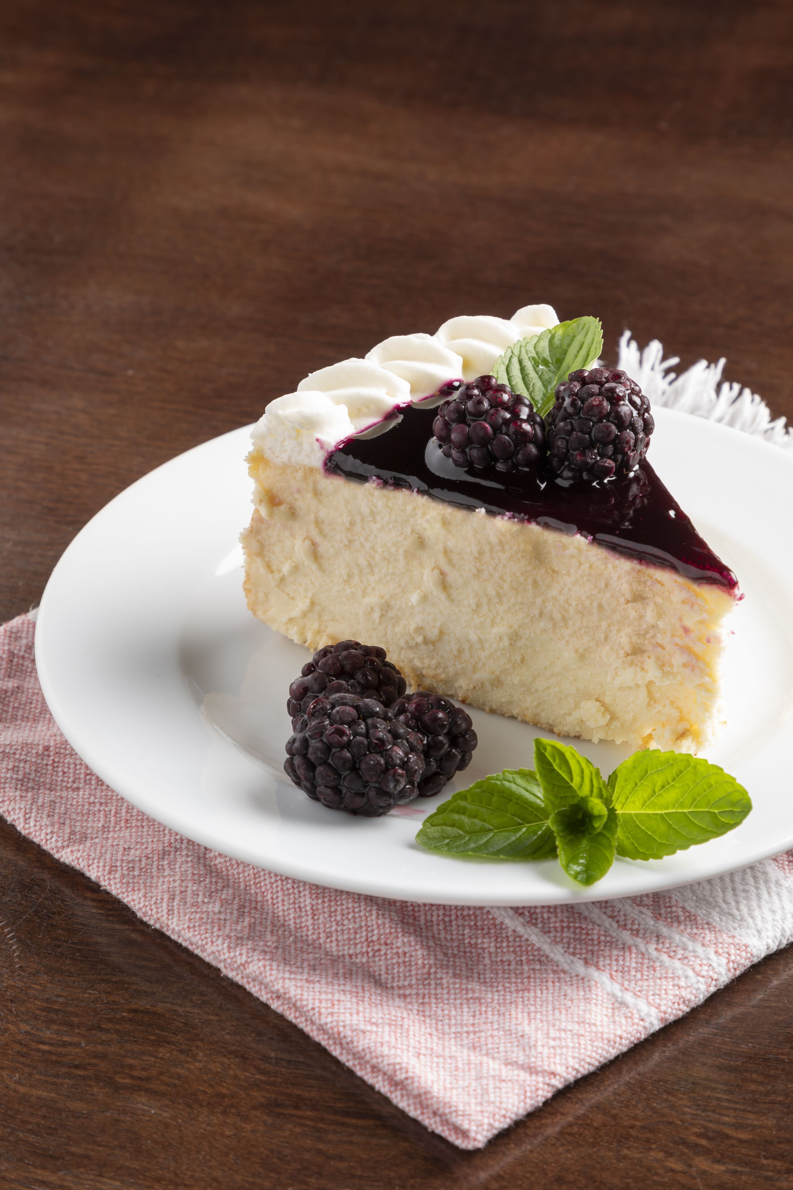 Una porción de pastel de queso con compota de frutos rojos sobre un plato blanco, acompañada de una hoja de menta y una baya entera.