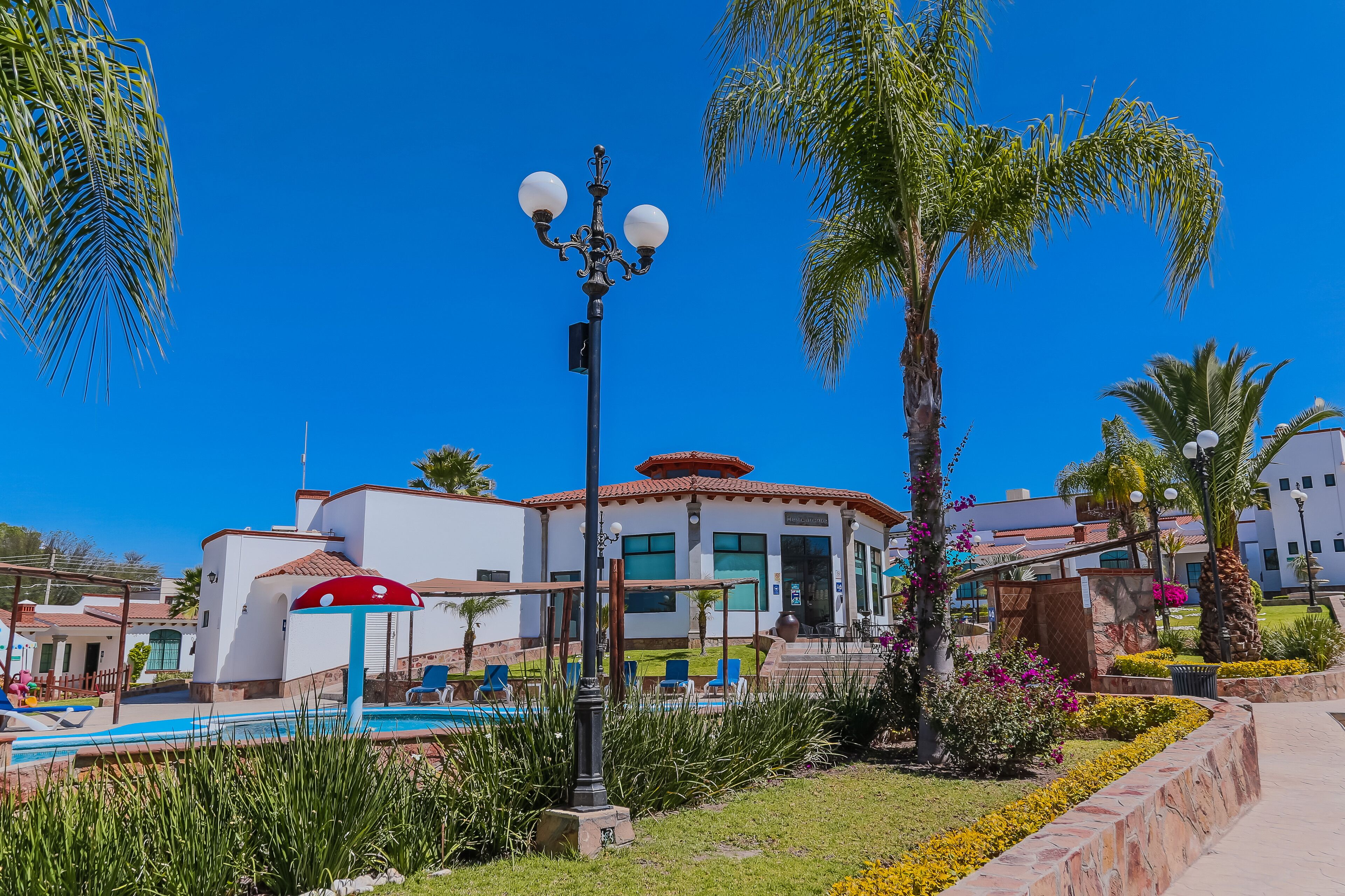 A pool area with lounge chairs, a lamp post, and a building with a red-tiled roof.