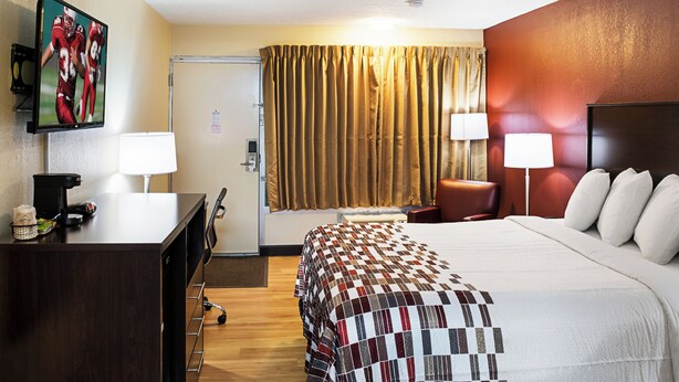 A hotel room with a bed, two bedside lamps, a desk with a coffee maker, a TV on the wall, and a window with curtains.