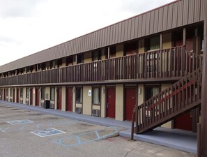 Un motel con una estructura de dos pisos, varias habitaciones con puertas rojas y un área de estacionamiento que incluye espacios para discapacitados.