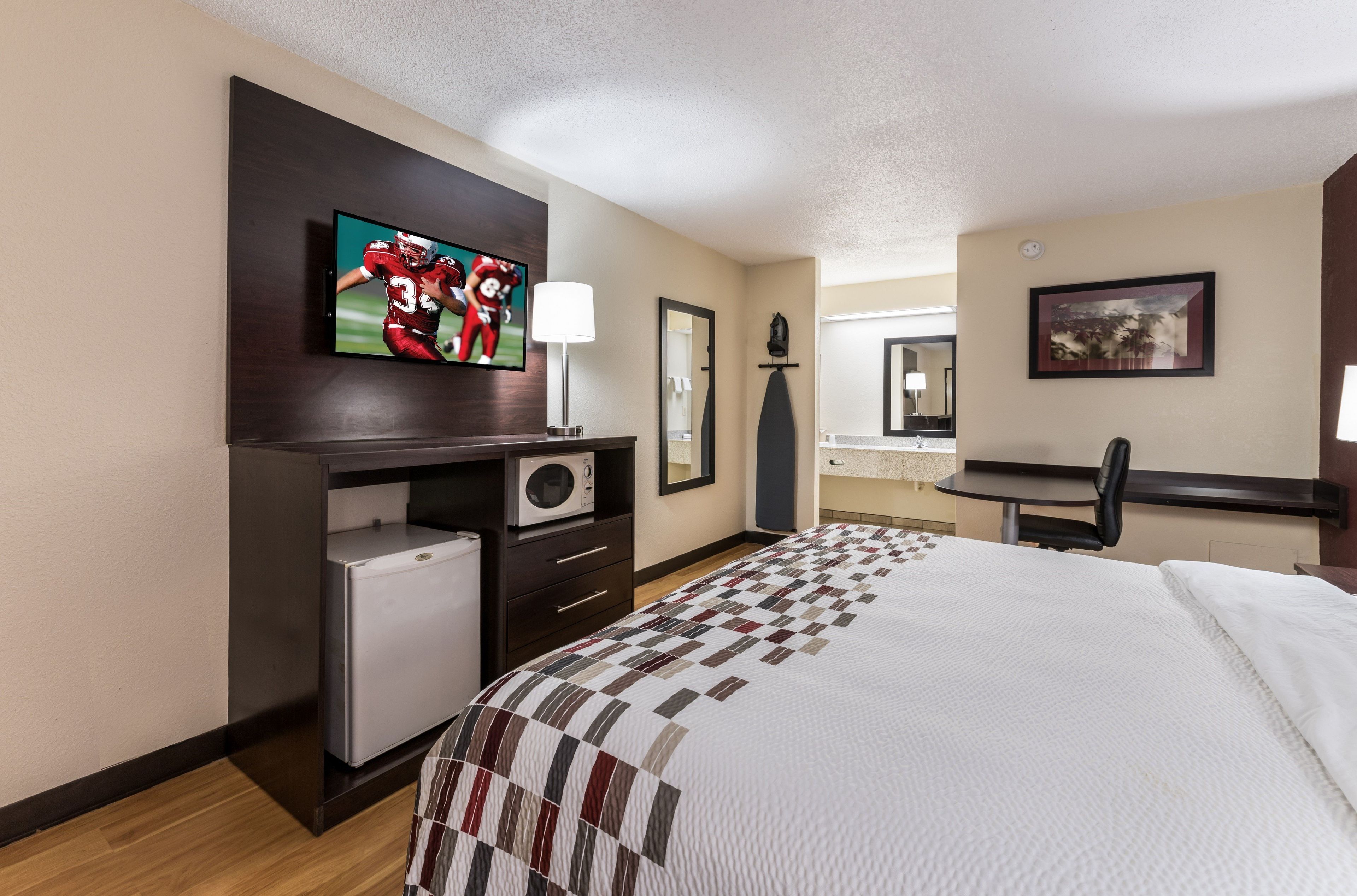 A hotel room with a bed, a television, a desk with a microwave, a chair, and a small refrigerator.