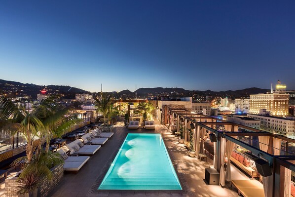 A rooftop pool with lounge chairs, a view of the cityscape, and a distant mountain range.