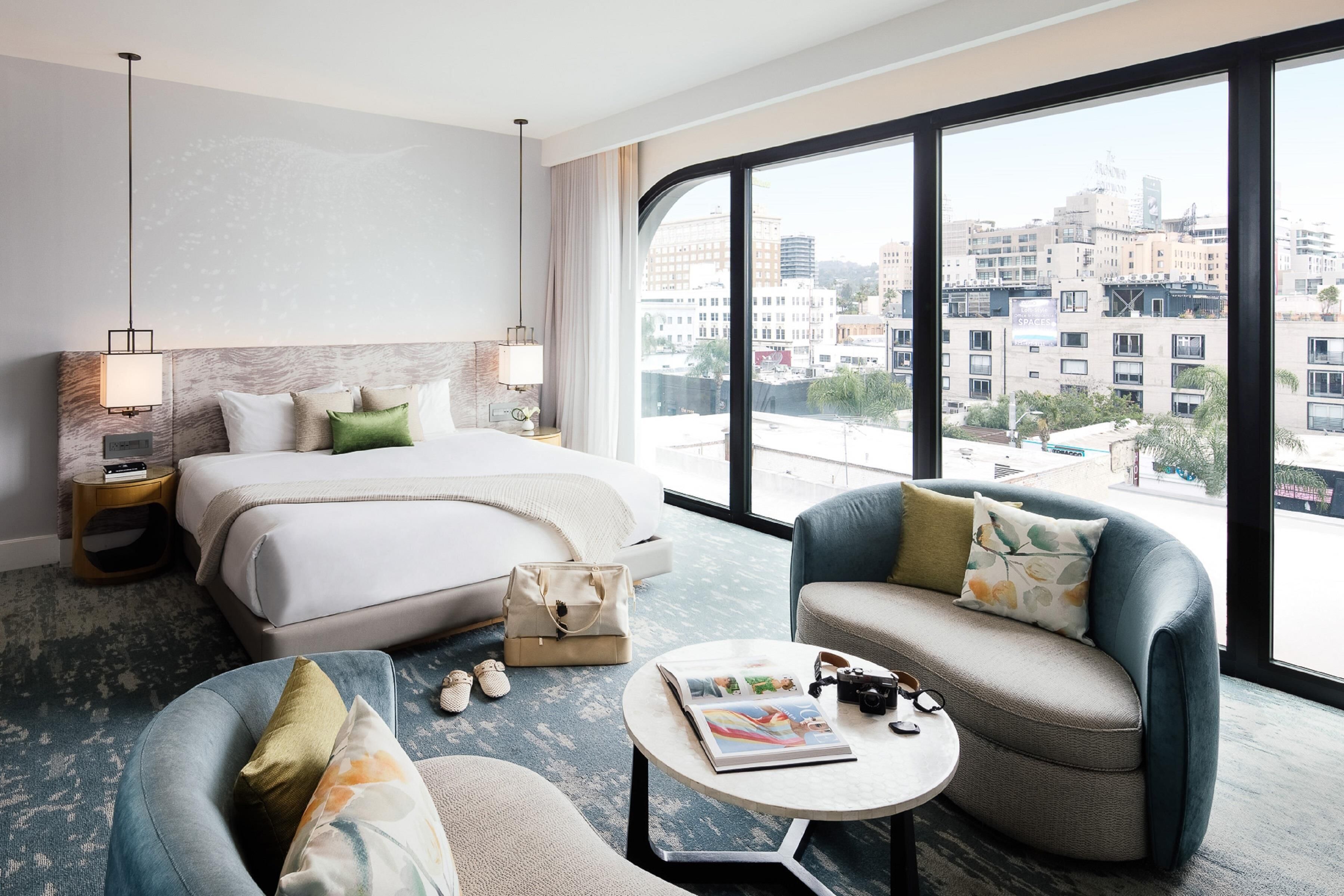 A hotel room with a bed, two armchairs, a coffee table, a view of the city through large windows, and a cityscape visible outside.