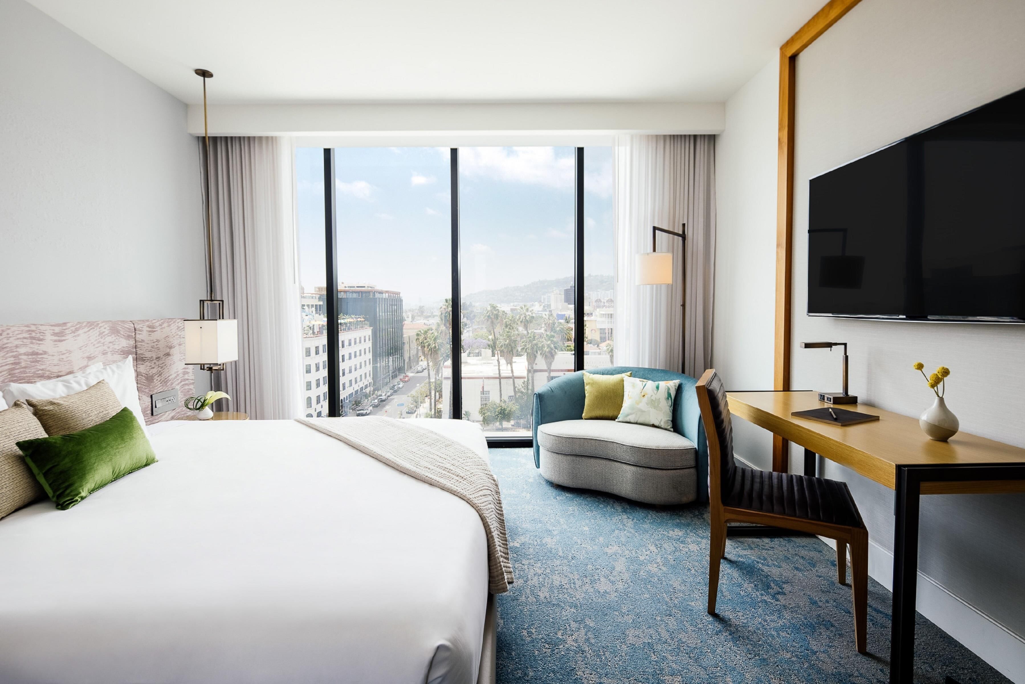 A hotel room with a large bed, a desk, a chair, a TV, and a view of the city.