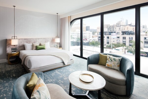 A hotel room with a bed, two armchairs, a small table, a large window, and a view of the cityscape.