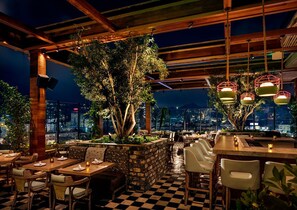 An outdoor dining area with checkered flooring, wooden tables, and chairs. There are hanging lights and a view of the cityscape in the background.