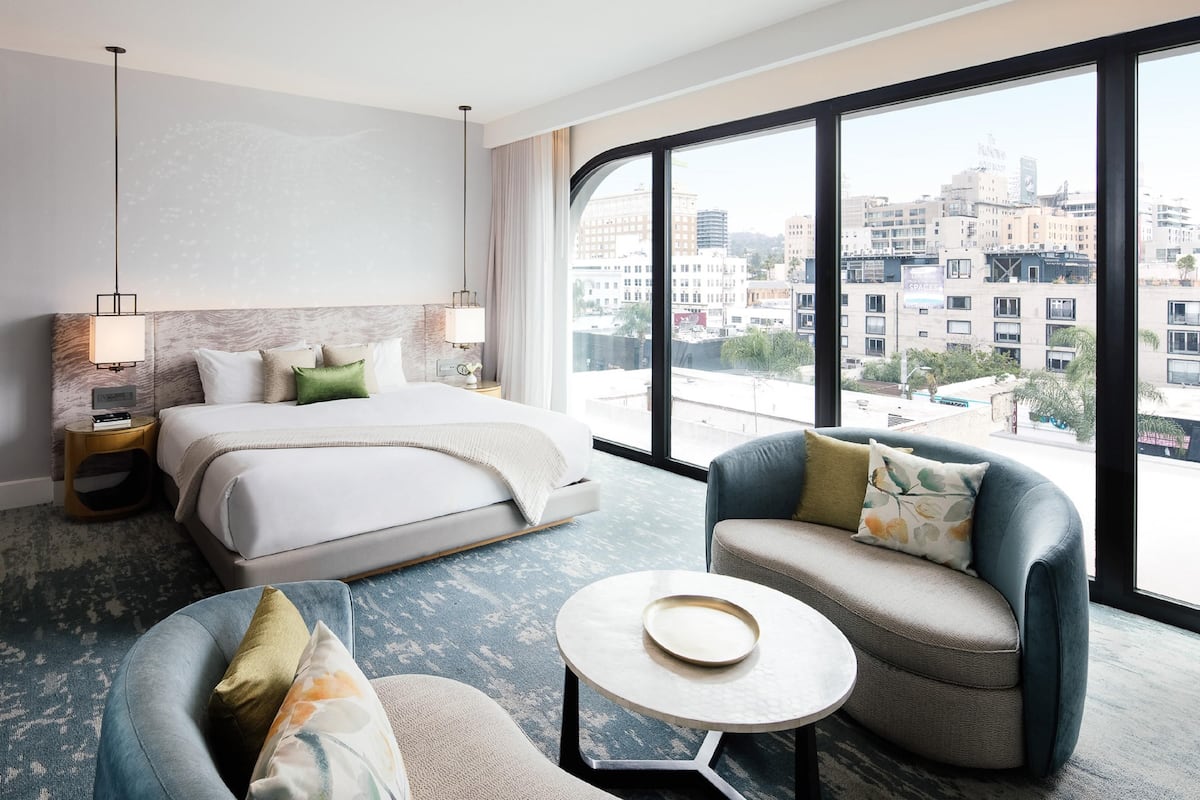 Une chambre d’hôtel avec un lit, deux fauteuils, une petite table, une grande fenêtre et une vue sur le paysage urbain.