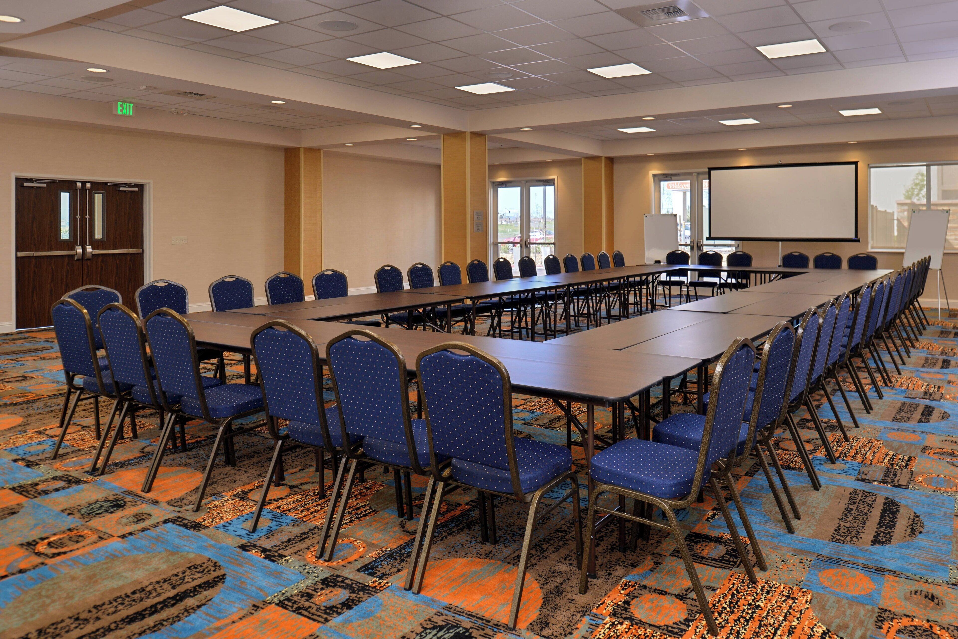 Une salle de conférence avec une longue table, des chaises bleues, un écran de projection et un sol recouvert de moquette.