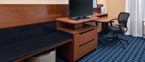 Une chambre d’hôtel avec un bureau en bois, une chaise noire, une télévision et une poubelle.