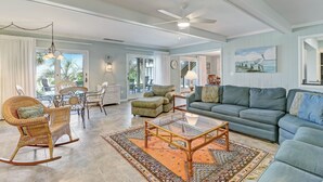 Un salón espacioso con un sofá grande, una mesa de centro de vidrio y un sillón mecedor de mimbre. Hay una zona de comedor con una mesa y sillas, y vistas al exterior a través de amplios ventanales.