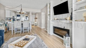 A modern living room with a fireplace, a TV mounted on the wall, and a dining area with a kitchen in the background.