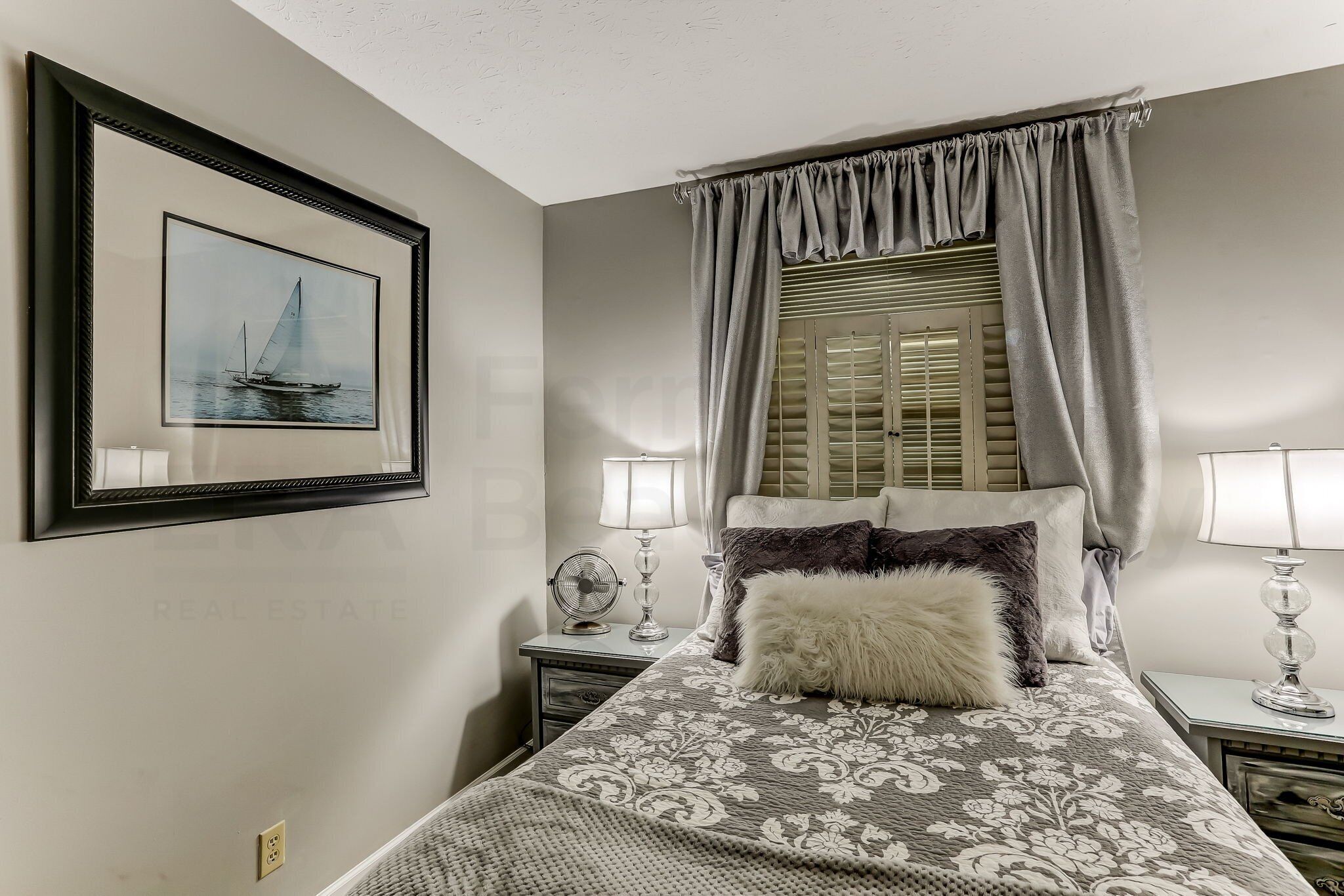 A bedroom with a bed, two bedside tables with lamps, a framed picture on the wall, and a window with curtains.