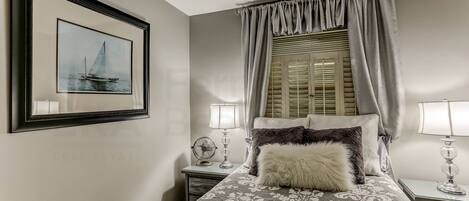 A bedroom with a bed, two bedside tables with lamps, a framed picture on the wall, and a window with curtains.