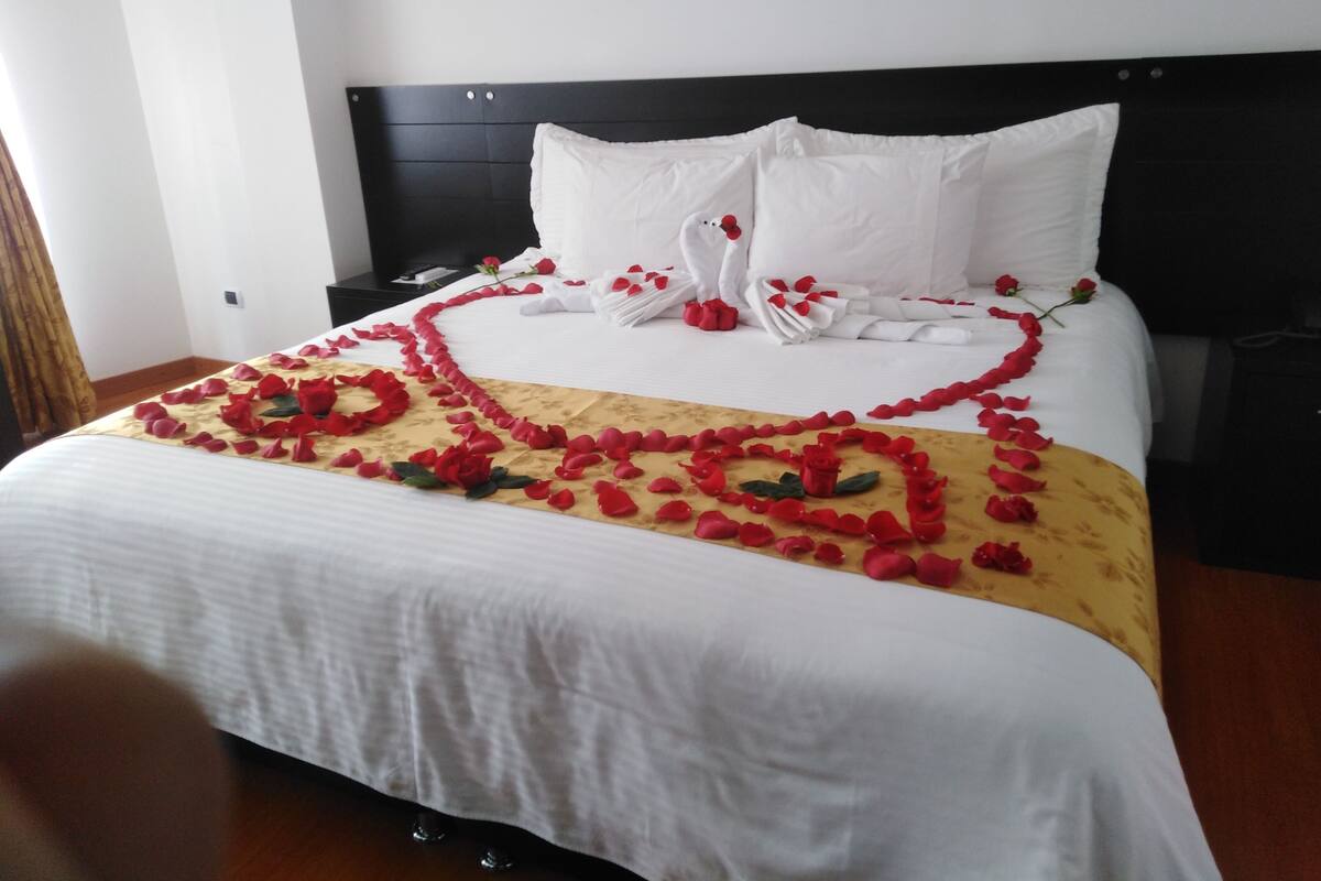 A neatly made bed with white linens, a black headboard, and red rose petals arranged in a heart shape on the bedspread.