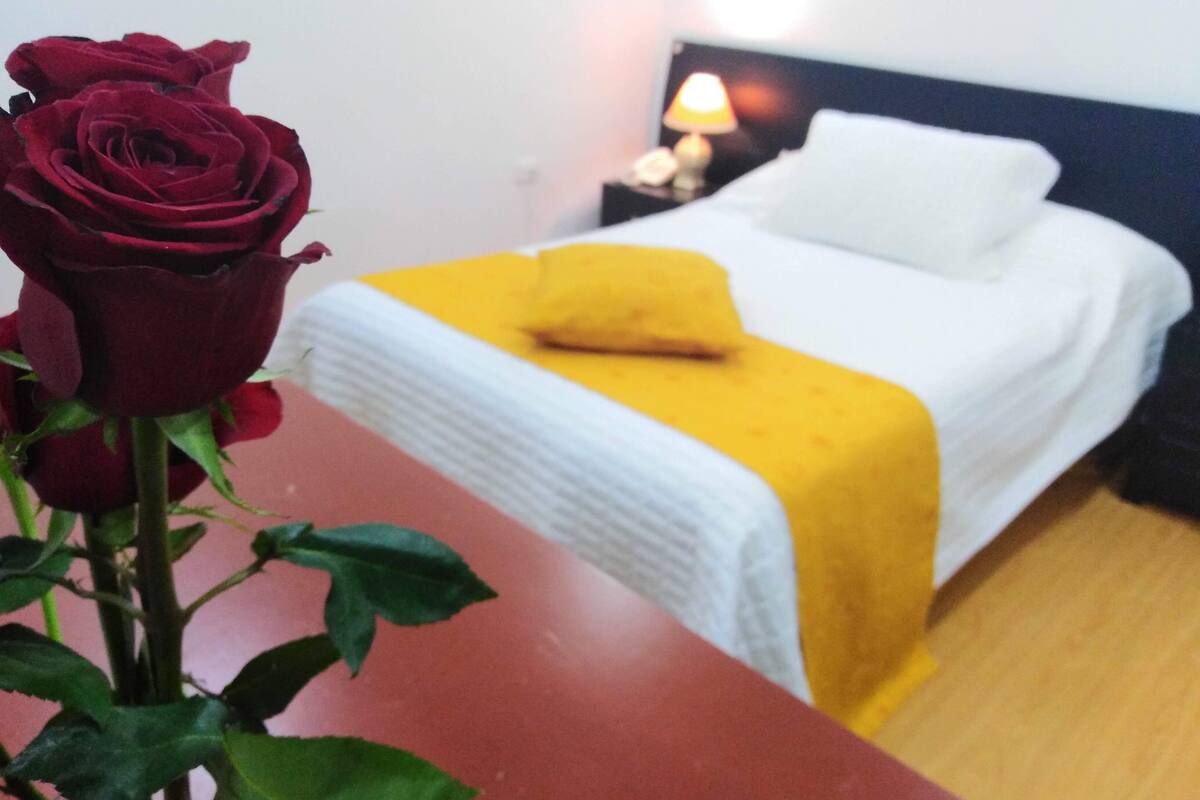 A hotel room with a bed, a nightstand, a lamp, and a red rose on a table.