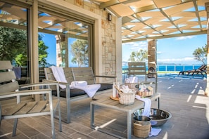 A sunlit outdoor seating area with stone walls, a view of the sea, and a table with amenities.
