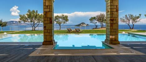 A pool area with a view of the sea, wooden flooring, and two lounge chairs under a canopy.