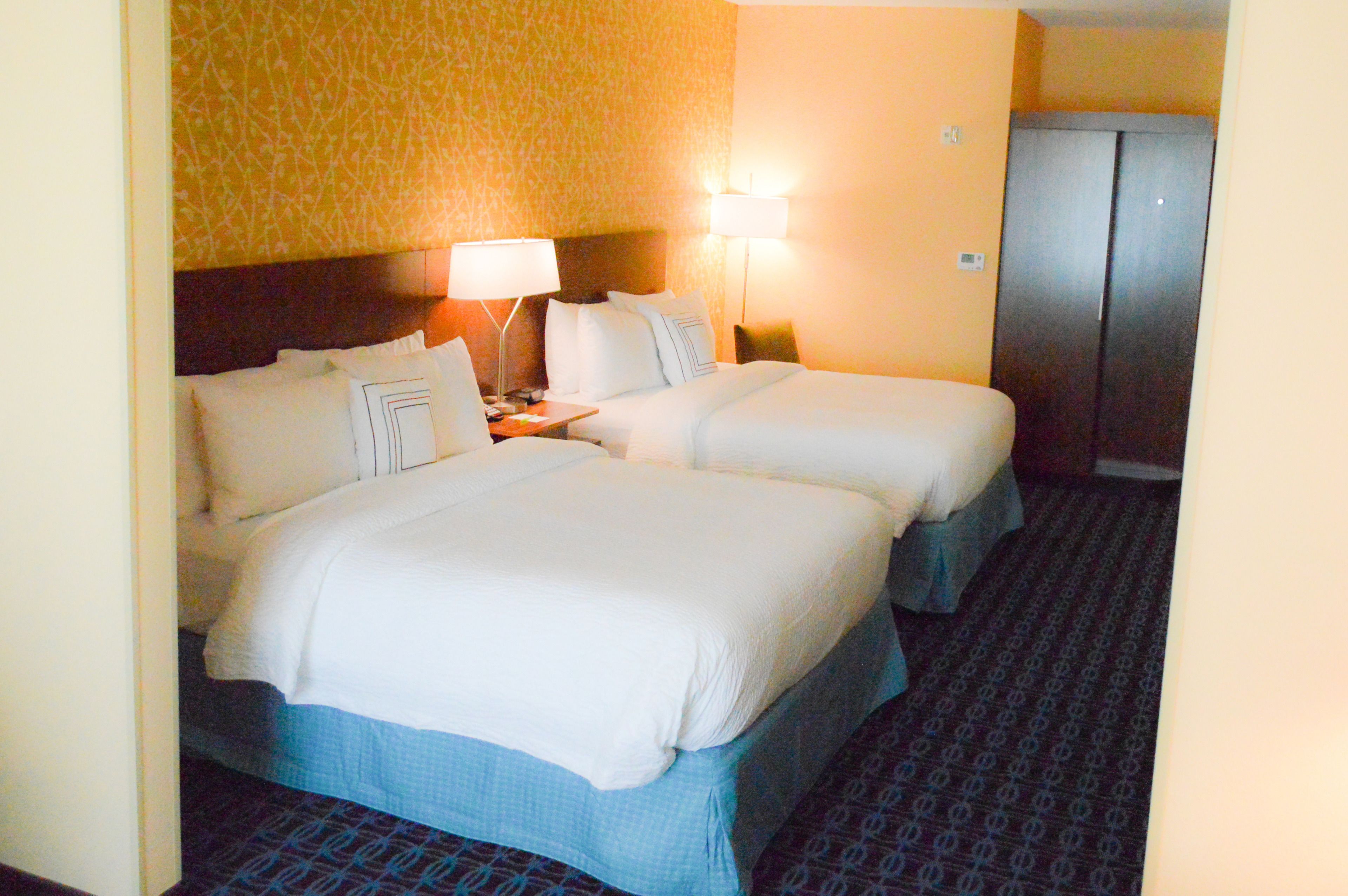 Quarto de hotel com duas camas, mesa de cabeceira, luminária e armário.