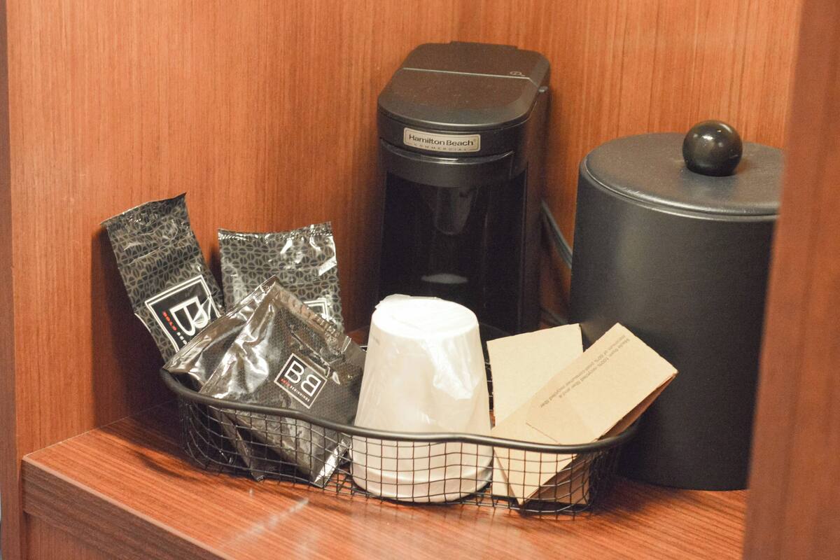 A hotel room's minibar with a coffee maker, a trash can, and a basket containing a coffee cup, a bag of snacks, and some papers.
