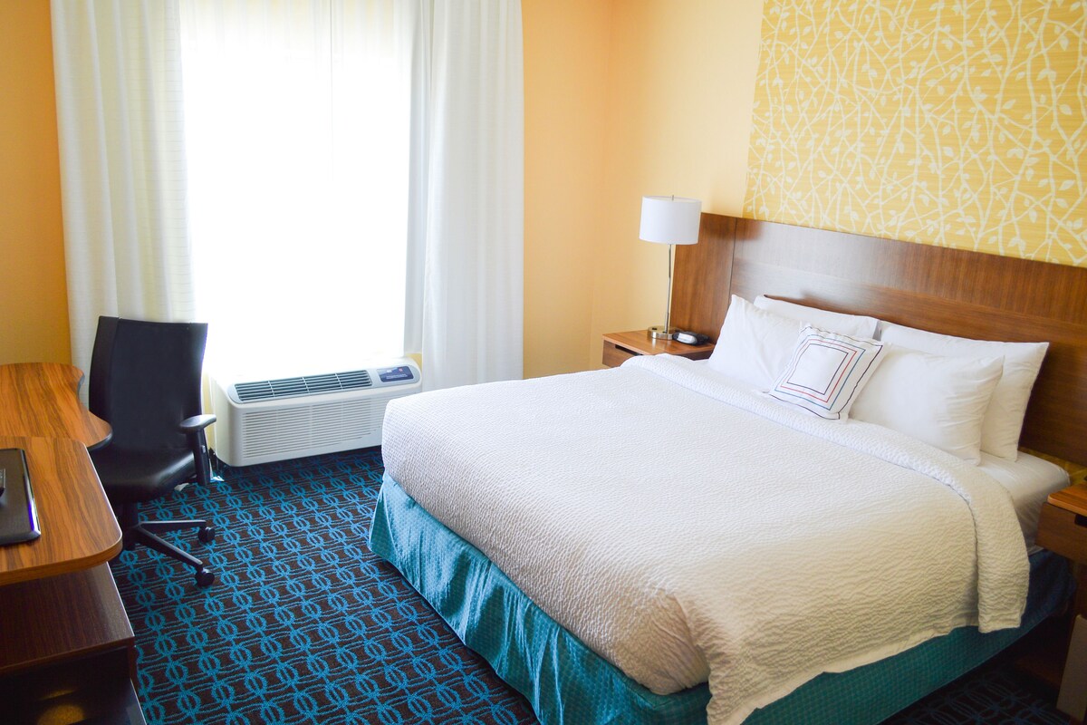 A hotel room with a bed, a desk, a chair, an air conditioning unit, and a window with curtains.