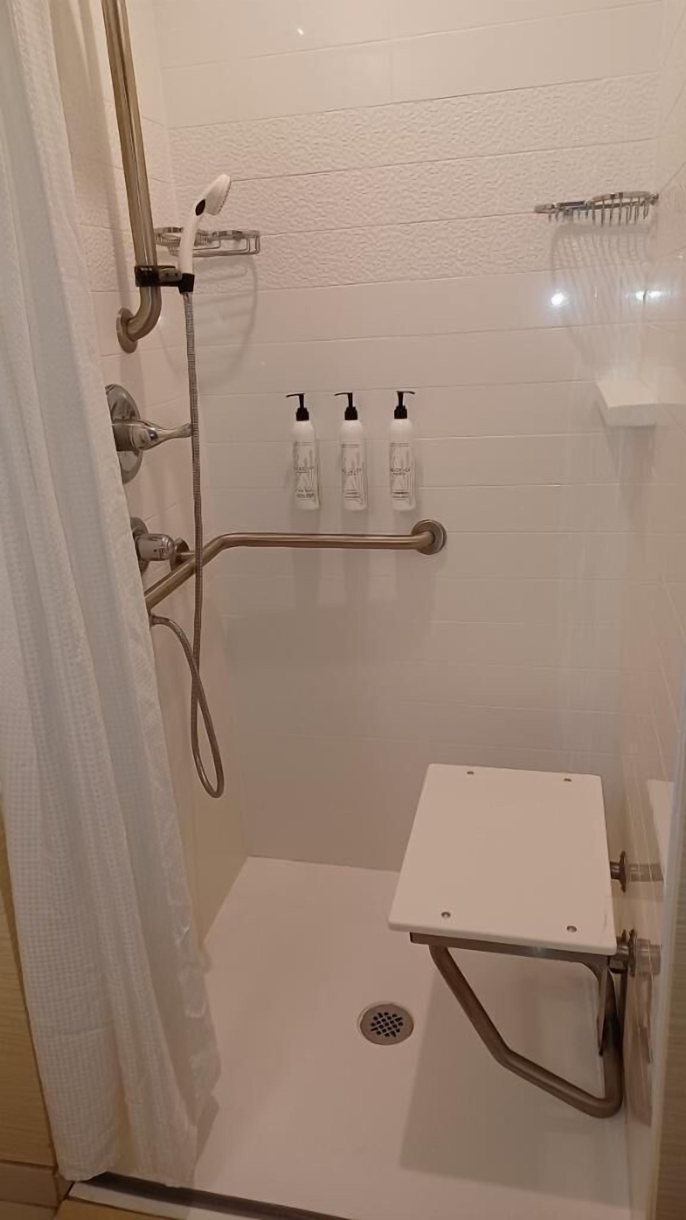 Ducha a ras de suelo con pared de azulejos blancos, cabezal de ducha manual, secador de mano y tres botellas de champú.