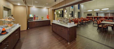 A hotel dining area with a buffet counter, wooden flooring, and a seating area with tables and chairs.