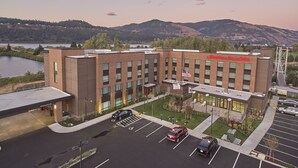 A modern hotel with a flat roof, a parking lot, and a view of a lake and mountains in the background. - Hampton Inn & Suites Hood River (Hood River)