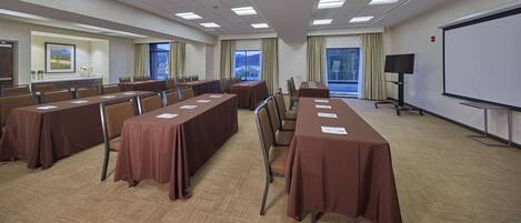 A conference room with long tables, chairs, a projector screen, and windows with curtains.