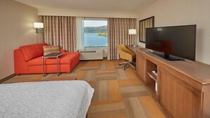 A hotel room with a bed, a red sofa, a desk with a television, and a view of a lake. - Hampton Inn & Suites Hood River (Hood River)