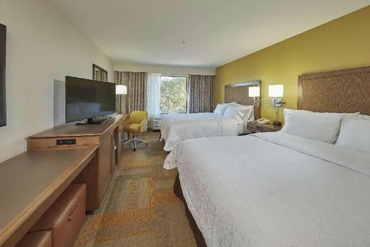 Habitación de hotel con dos camas, un escritorio con televisor, una silla y ventana con cortinas.