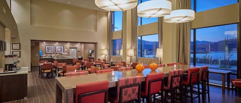 A spacious hotel lobby with a long communal table, red upholstered chairs, and large windows offering a view of the outdoors.