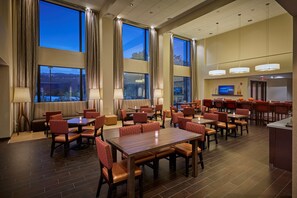 Une salle à manger d’hôtel avec de nombreuses tables et chaises, de grandes fenêtres avec des rideaux et un téléviseur fixé au mur.