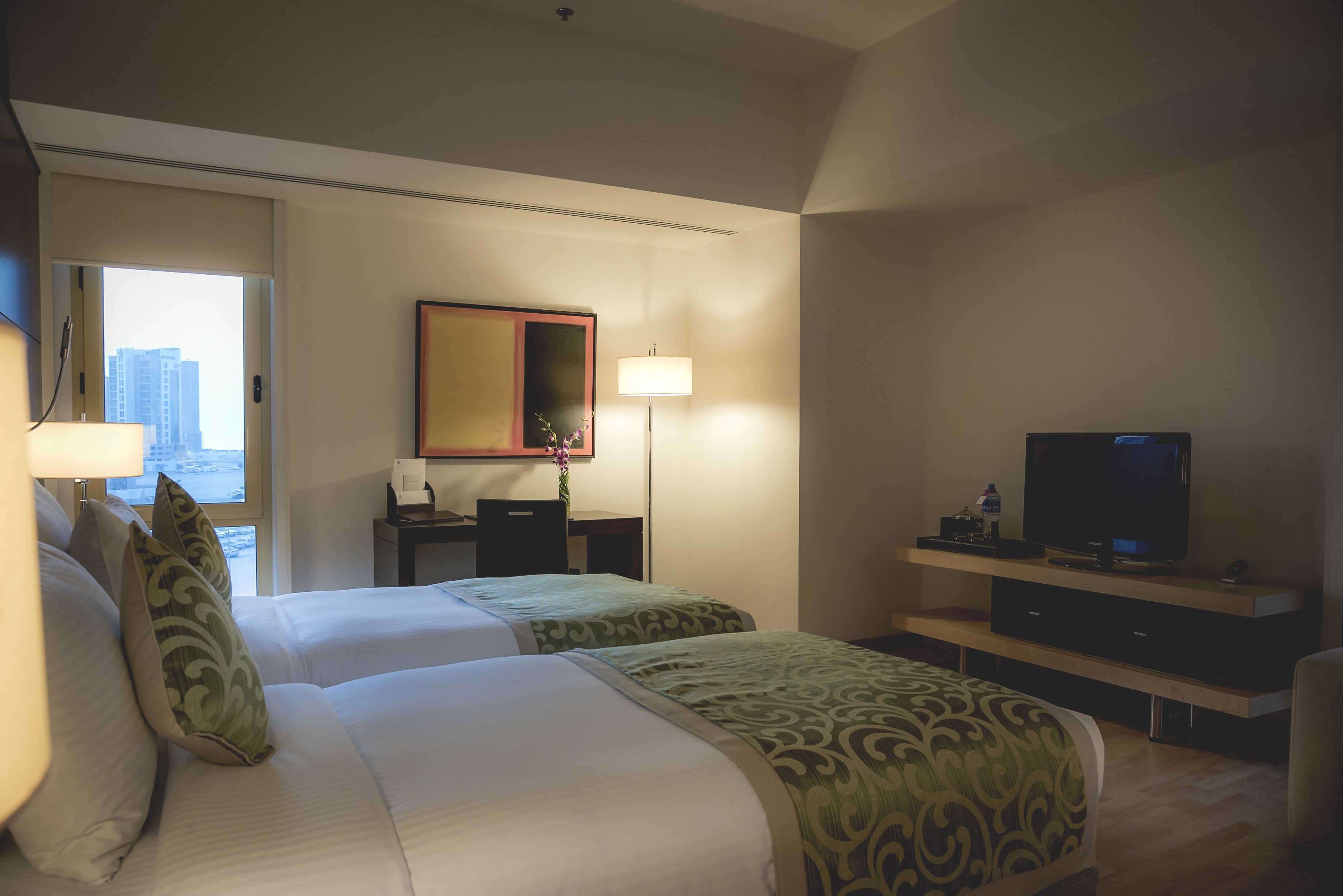 A hotel room with two beds, a desk, a TV, and a window with a city view.