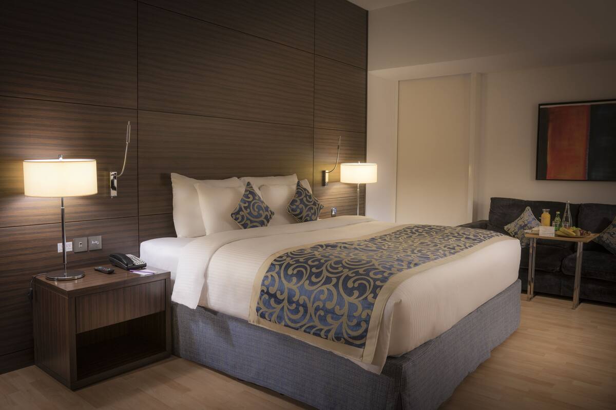 A hotel room with a large bed, two bedside lamps, a nightstand with a phone, a small table with a bottle and fruit, and a dark-colored sofa.